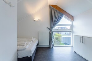 Une chambre à coucher avec un lit, un radiateur, une fenêtre avec des rideaux et une vue sur l’extérieur.