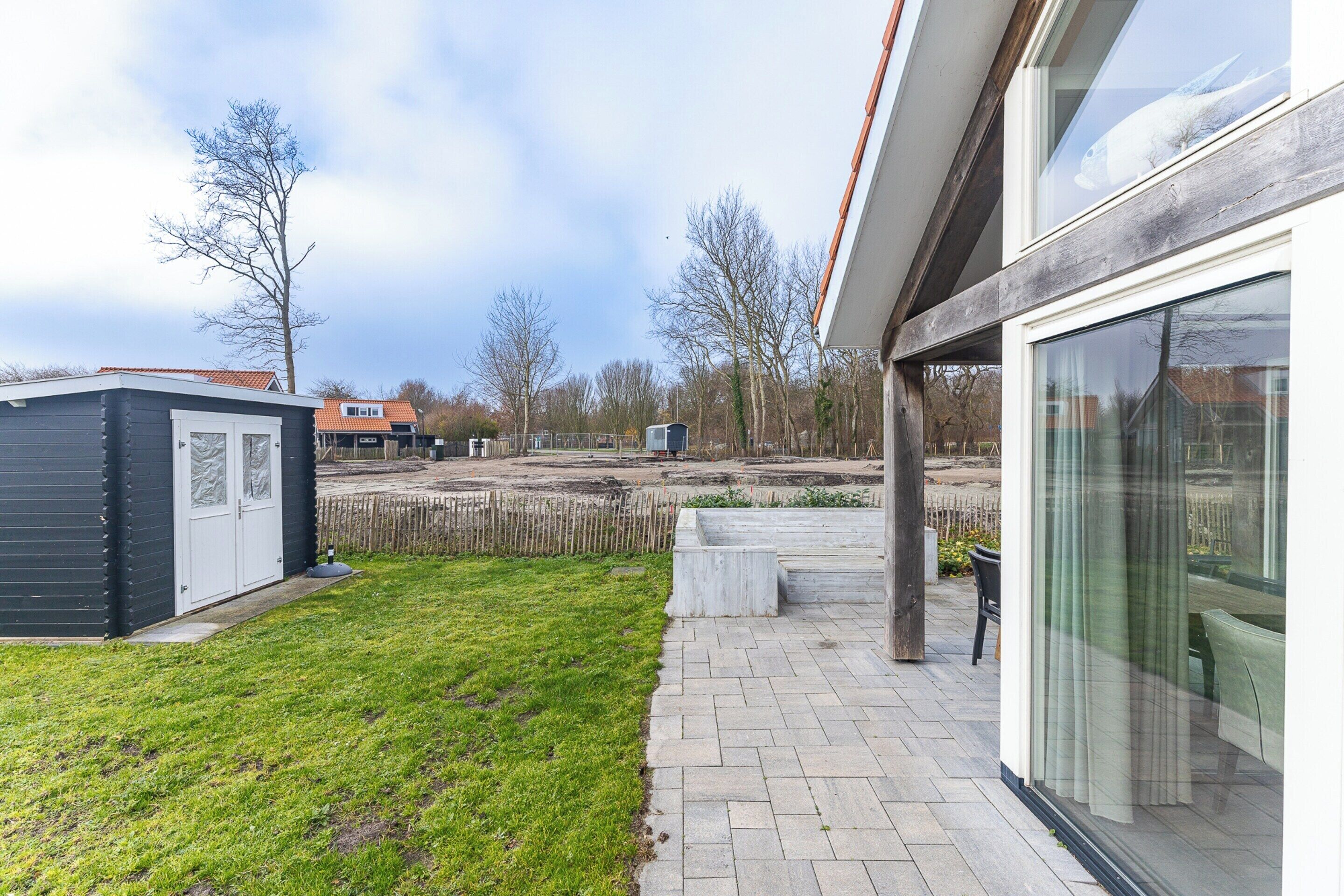 Ein modernes Haus mit Pflasterterrasse, kleiner Gartenhütte und großem Fenster mit Blick auf einen Garten.