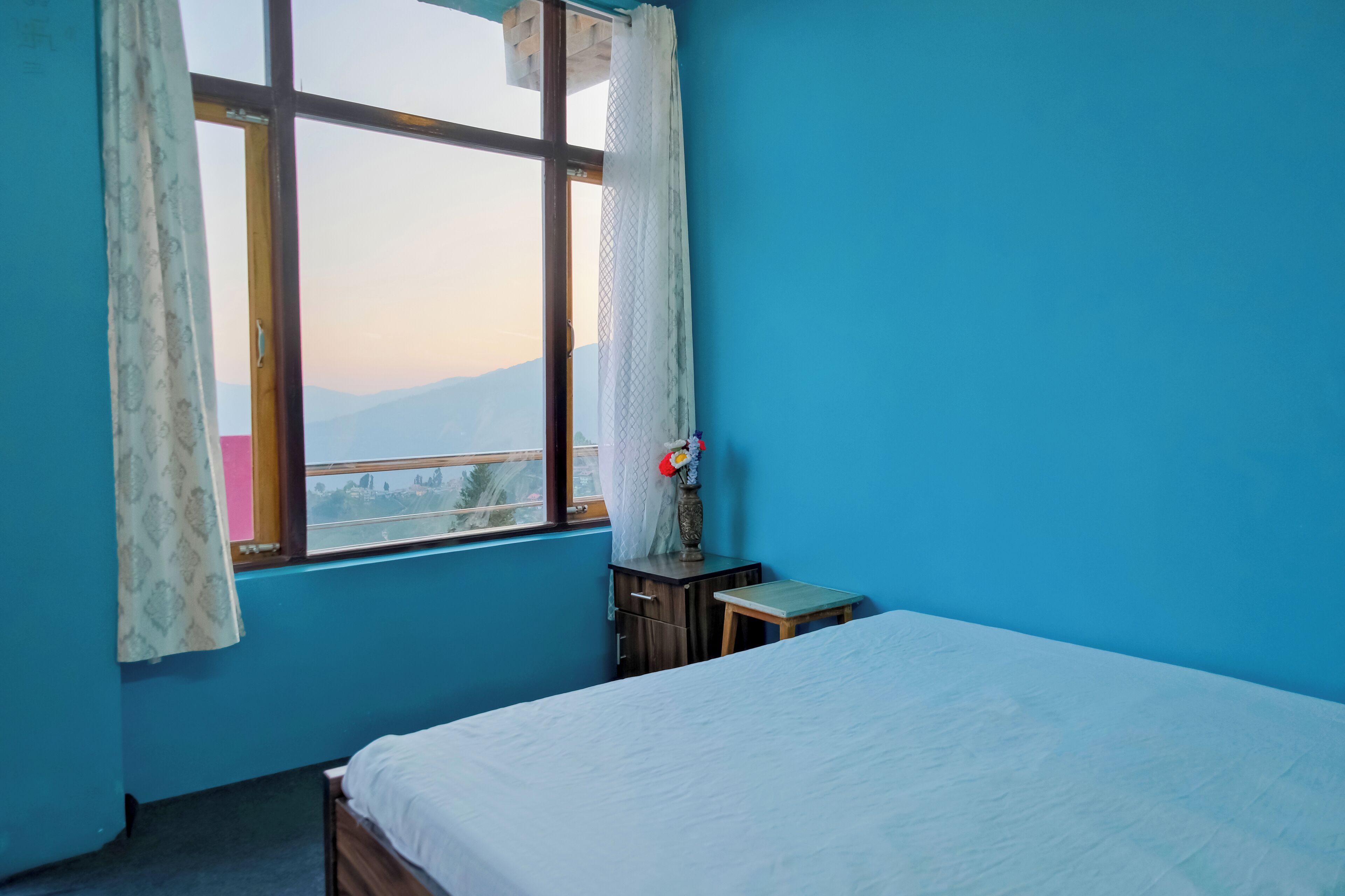 A bedroom with a bed, a window with curtains, a small table, and a nightstand.