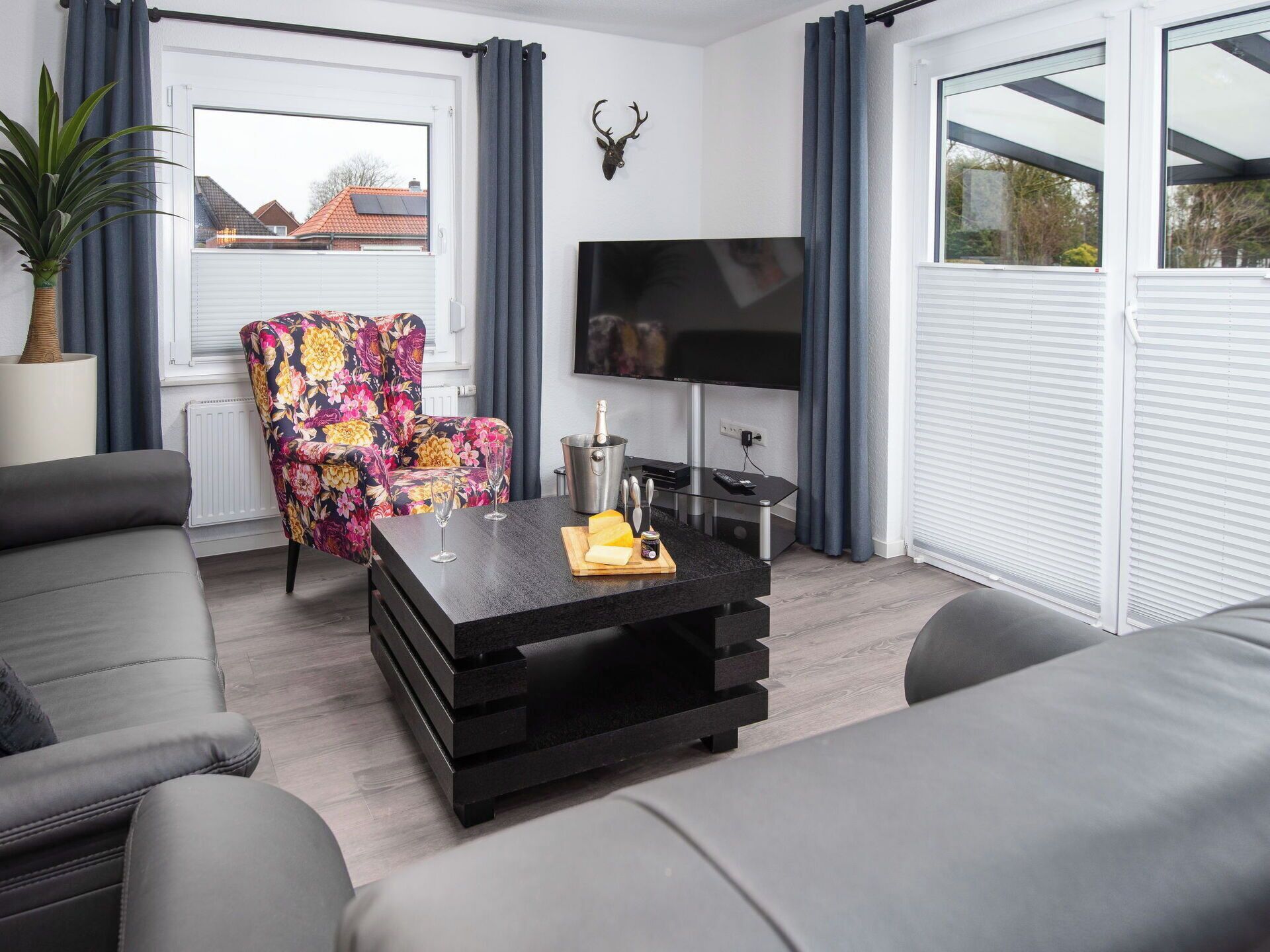 Un salon moderne comprenant un canapé, un fauteuil à motifs floraux, une table basse avec une bouteille et des verres, un téléviseur à écran plat et une décoration représentant une tête de cerf.