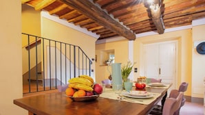 A dining area with a wooden table set for four, surrounded by purple chairs. A bowl of fruit is on the table, and there is a pitcher and a vase with flowers. - Beautiful private villa for 10 guests with private pool, WIFI, TV, balcony and pets allowed (Capannori)