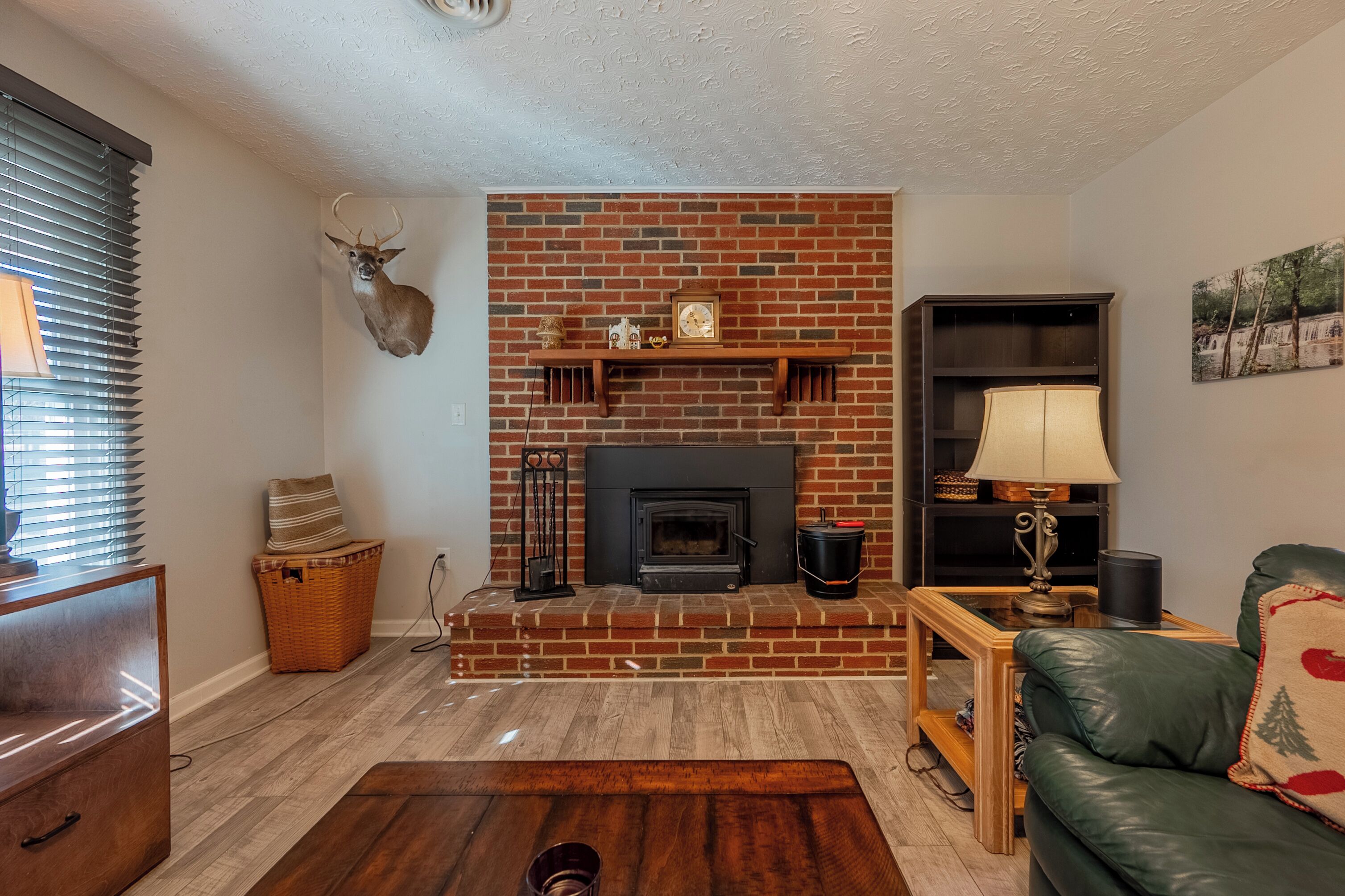 A living room with a brick fireplace, a mounted deer head, a green leather sofa, and a wooden coffee table.