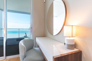 Ein modernes Hotelzimmer mit großem Fenster und Meerblick, einem runden Spiegel, einem Nachttisch mit Lampe und einem bequemen Sessel.