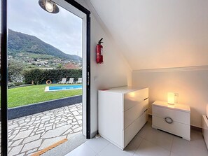 A modern bedroom with a view of a swimming pool and mountains. It includes a white wardrobe, a bedside table with a lamp, and a fire extinguisher on the wall.