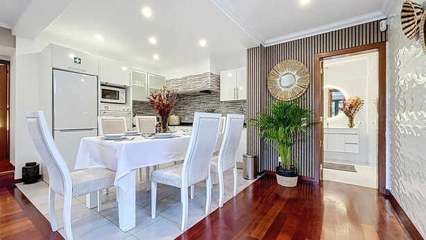 Une cuisine moderne avec des meubles blancs, un coin repas avec une table et des chaises blanches, et une plante en pot.