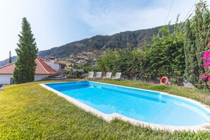 Ein Swimmingpool mit klarem, blauem Wasser, umgeben von Gras, Bäumen und Blick auf eine Hügelside mit Häusern.