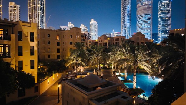 Vista noturna de uma área residencial com edifícios modernos, piscina e palmeiras.