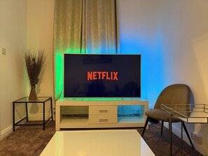 En stue med et fladskærms-tv på et hvidt tv-møbel, en vase med tørrede planter og en stol.