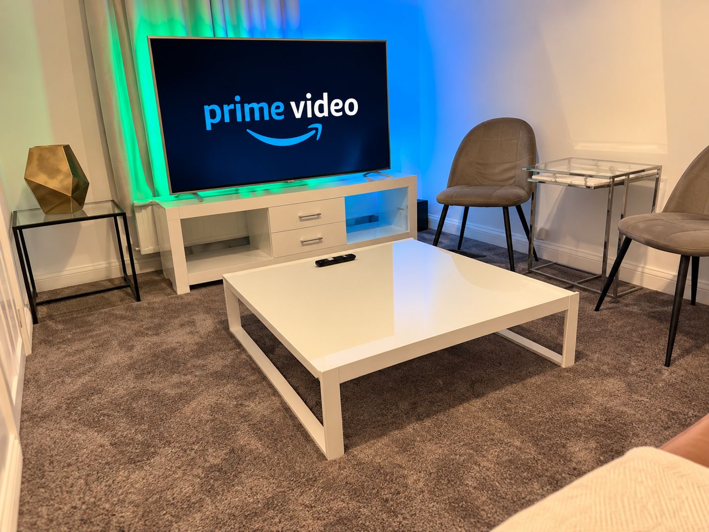 A living room with a flat-screen TV displaying 'prime video' on the screen, a white coffee table, two chairs, and a glass side table with a decorative object.