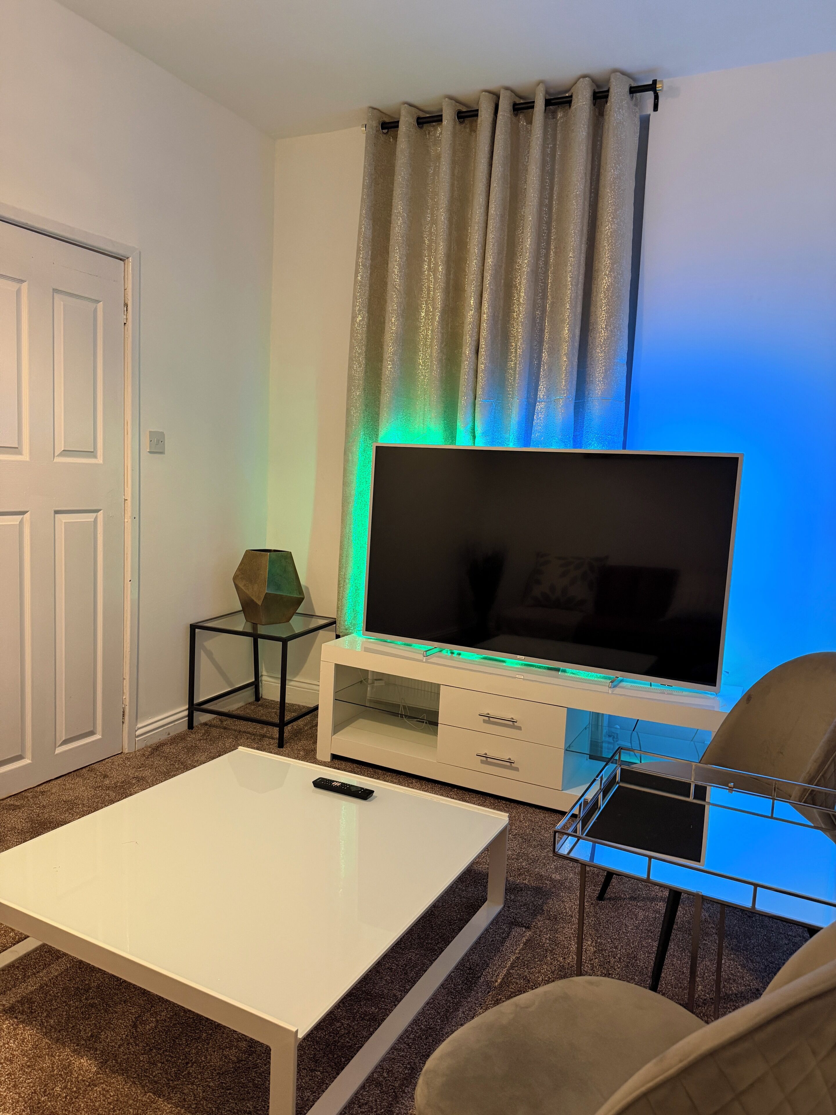 Un salon avec un téléviseur à écran plat posé sur un meuble de télévision blanc, une table basse en verre, un fauteuil beige et une porte sur le côté gauche.