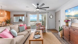 A living room with a sofa, coffee table, and a chair. There is a ceiling fan, a window with curtains, and a view of the ocean. - Flamingo Cove  | by Pitzer-Properties (Port St. Joe)