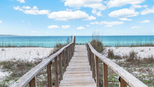 A wooden boardwalk leading to a beach with clear blue water and a partly cloudy sky. - Flamingo Cove  | by Pitzer-Properties (Port St. Joe)