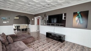 A living room with a large TV, a sofa, and a dining area in the background.