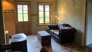Une pièce rustique avec un canapé en cuir, une malle en bois et une petite table. Il y a deux fenêtres et une porte.