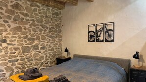 Une chambre avec un mur en pierre, un lit recouvert d’une couverture bleue, deux oreillers jaunes et trois œuvres d’art représentant des bicyclettes, accrochées au mur.