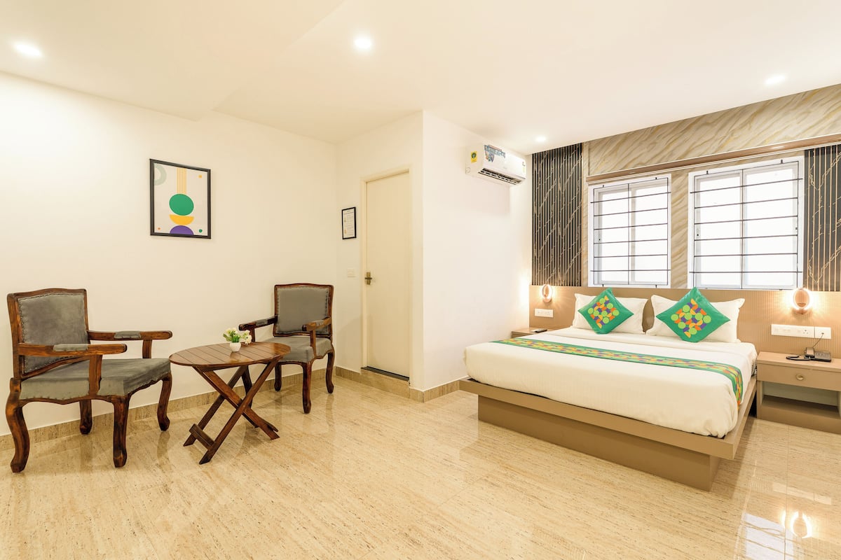 Una habitación de hotel con una cama, dos sillas de madera, una mesita, una mesita de noche y un ventanal con cortinas.