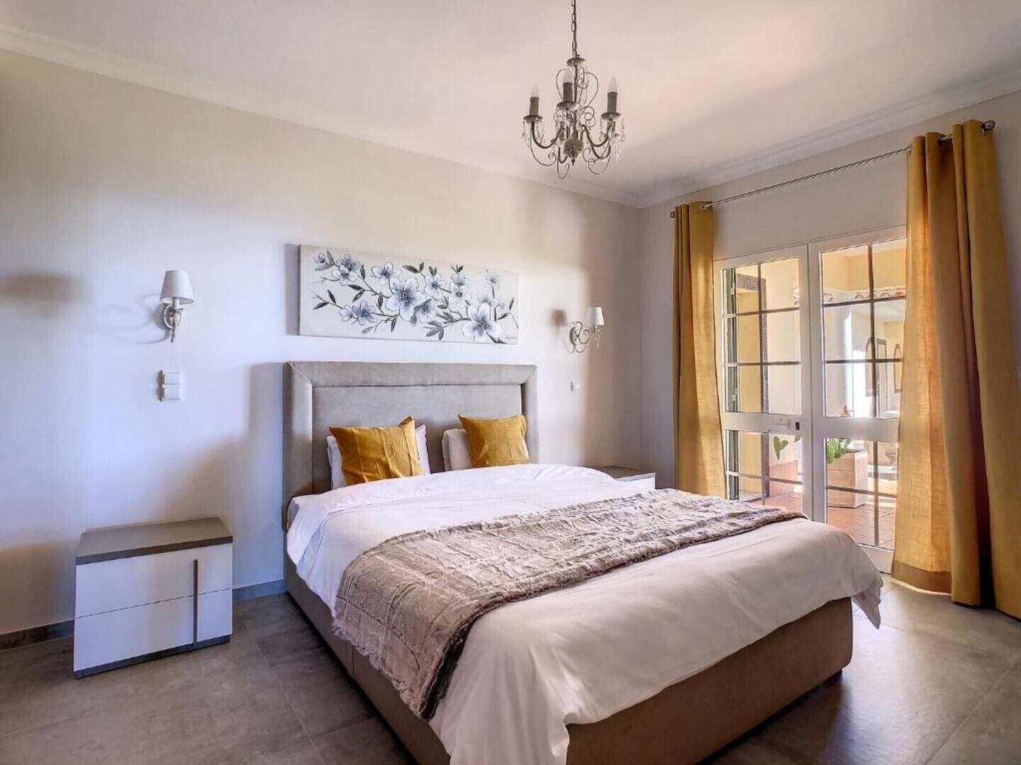 A bedroom with a large bed, a nightstand, a chandelier, a wall-mounted lamp, and a window with curtains.