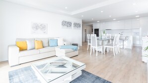 Egy modern nappali, melyben fehér kanapé, üveg kávézóasztal és fehér székekkel, valamint üveg asztallal berendezett étkező található.
