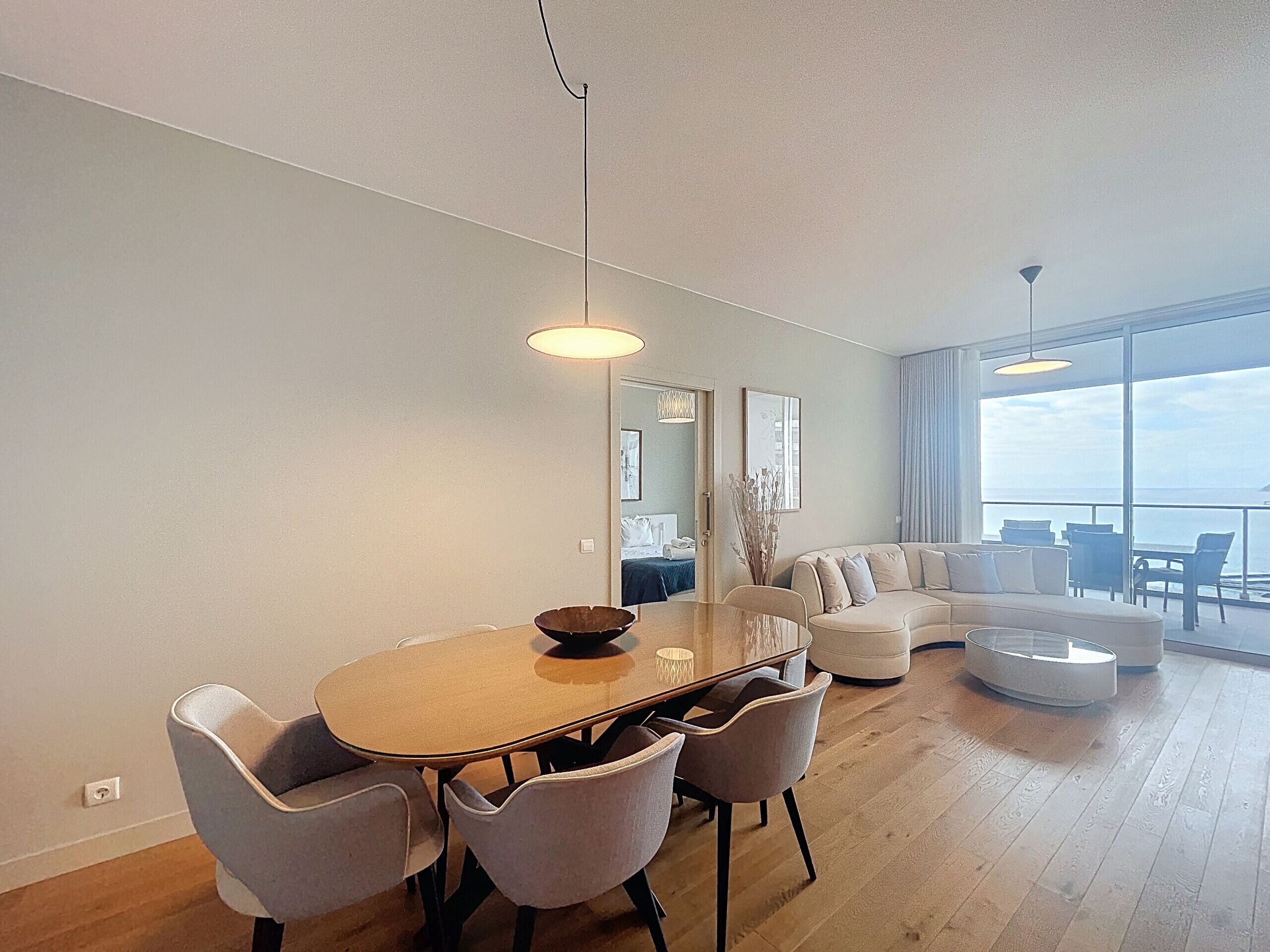 Un salon moderne avec une table à manger ronde, des chaises recouvertes d’un tissu gris, un canapé et une grande fenêtre donnant sur la mer.