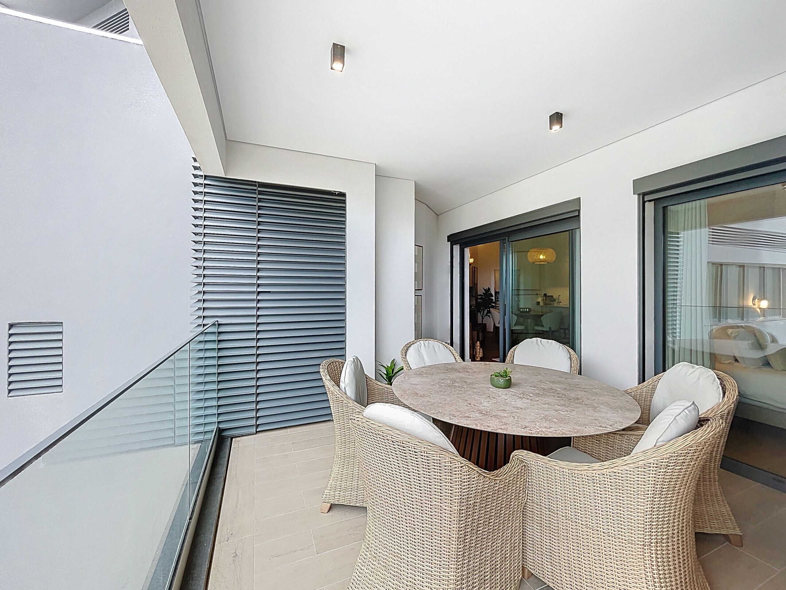 A modern balcony with a round table and wicker chairs, featuring a glass door leading to an indoor area.