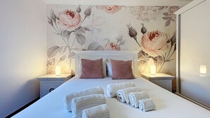 En pænt redt seng med sammenrullet håndklæder, to sengeborde med lamper, og blomstret tapet.