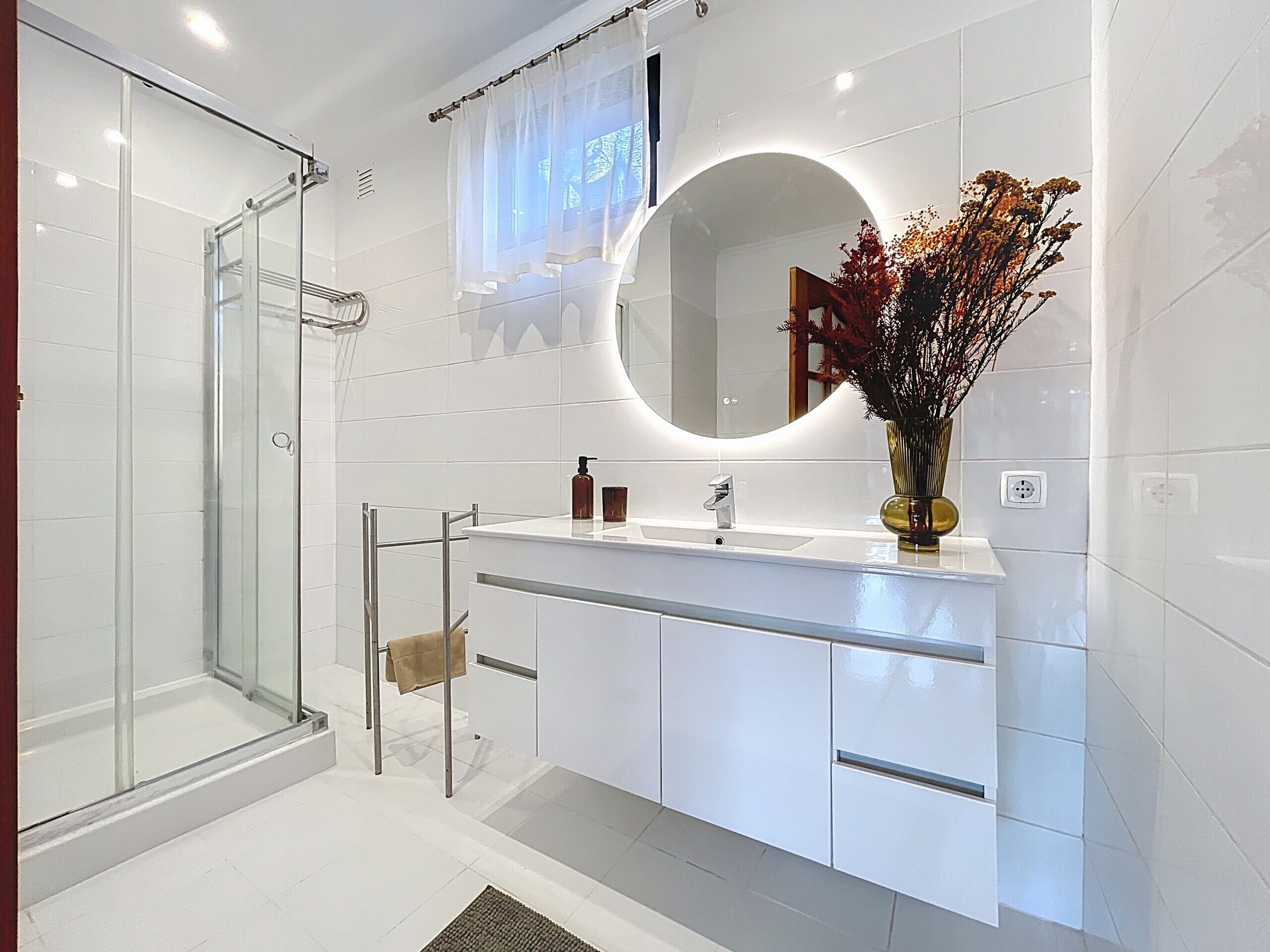 A modern bathroom with a glass shower enclosure, a white vanity with a round mirror, and a vase with dried flowers.