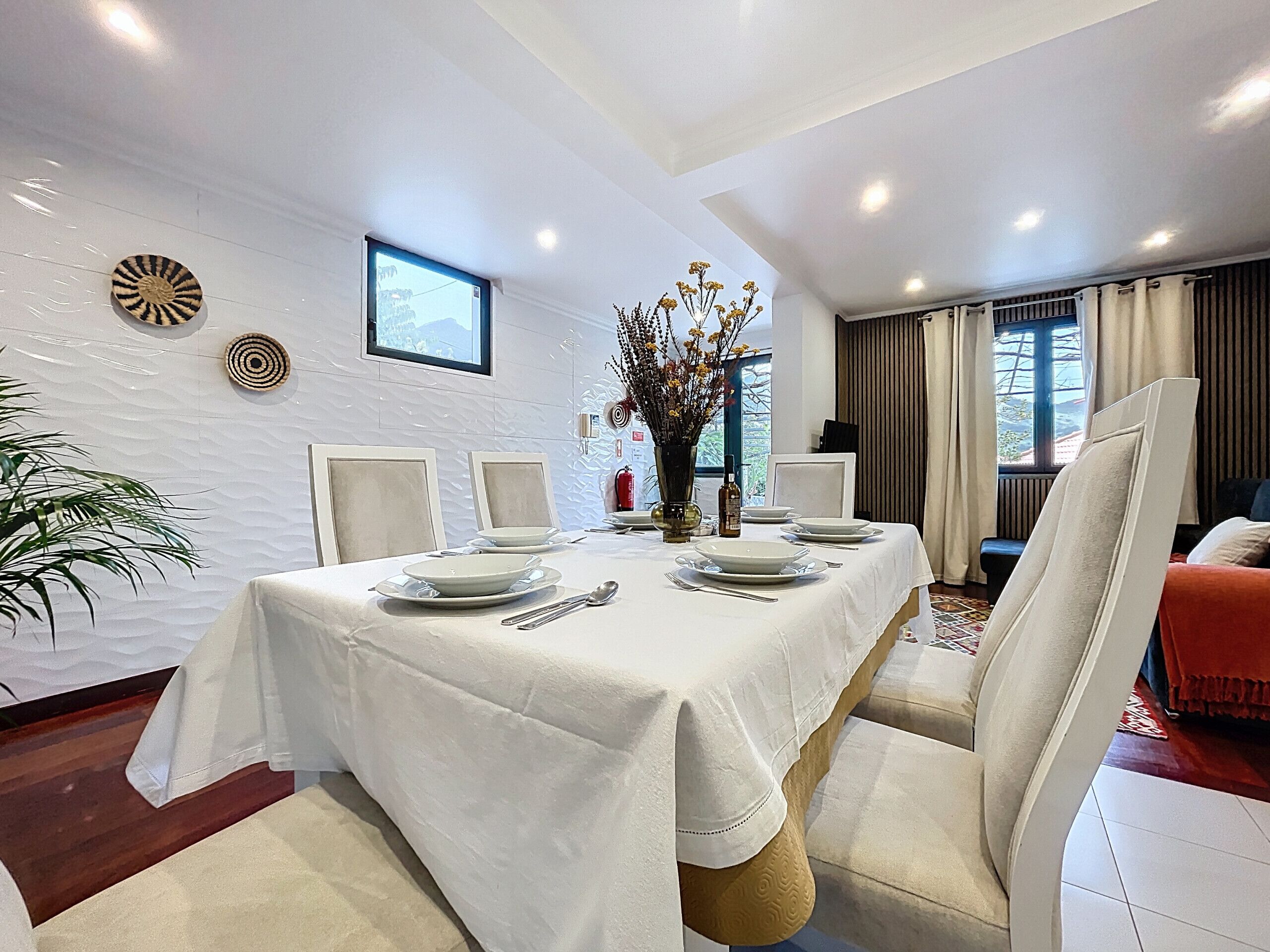 A dining area with a white table set for four, surrounded by white chairs. There is a vase with flowers on the table, and a window with curtains in the background.