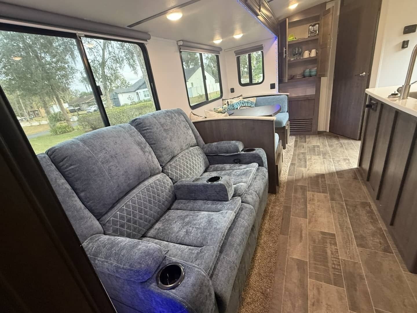 A compact RV interior with a gray sofa, a kitchen area, and large windows offering a view of the outdoors.