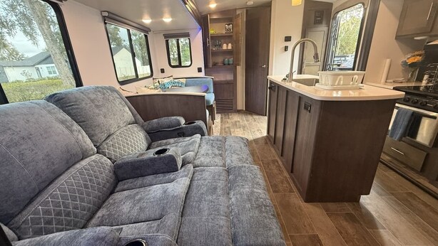 A compact RV interior with a kitchenette, a dining area, and a seating area with cushioned recliners.