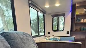 A row of grey upholstered seating with cup holders, a kitchen area with a sink and cabinets, and large windows with a view of trees.