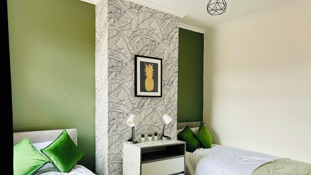 Un dormitorio con dos camas, almohadas verdes, una mesita de noche con lámparas, un cuadro con una piña y un papel tapiz estampado.