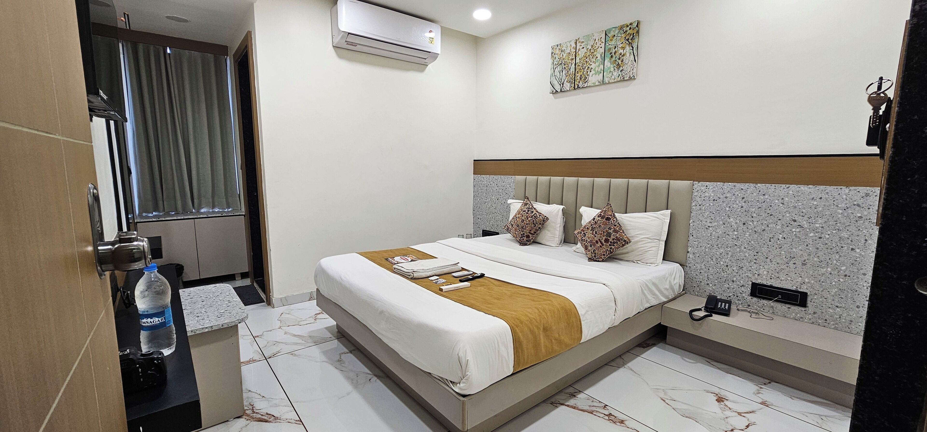 A hotel room with a bed, a bedside table, an air conditioner, a window with curtains, and a wall-mounted artwork.