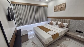 A modern hotel room with a large bed, a flat-screen TV mounted on the wall, and a window with vertical blinds. - Hotel Rose Gold (Ahmedabad)