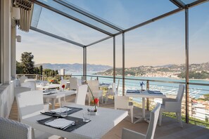 A restaurant with white wicker furniture, a glass enclosure, and a view of the sea and mountains. - Europa Grand Hotel  (Lerici)
