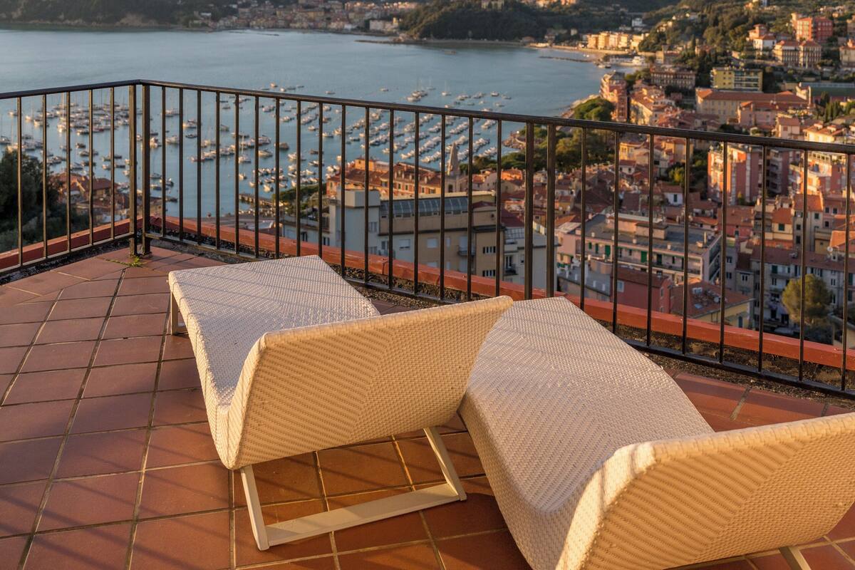 A terrace with two wicker lounge chairs overlooking a marina and a coastal town.