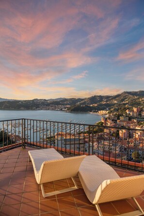 A terrace with two wicker lounge chairs overlooking a marina and a coastal town. - Europa Grand Hotel  (Lerici)