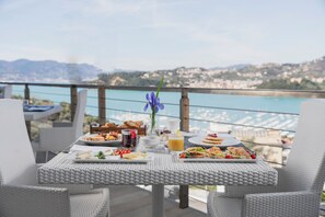 A dining table set on a balcony with a scenic view of a coastal town and mountains in the background. - Europa Grand Hotel  (Lerici)