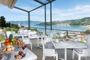 A terrace with white wicker furniture, a fruit basket, and a view of a marina and coastal town. - Europa Grand Hotel  (Lerici)