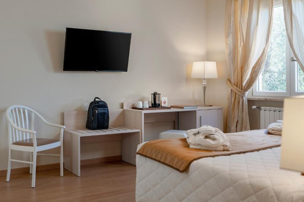 Una camera d'albergo con un letto, una scrivania, una sedia, una televisione, una lampada e una finestra con tende.