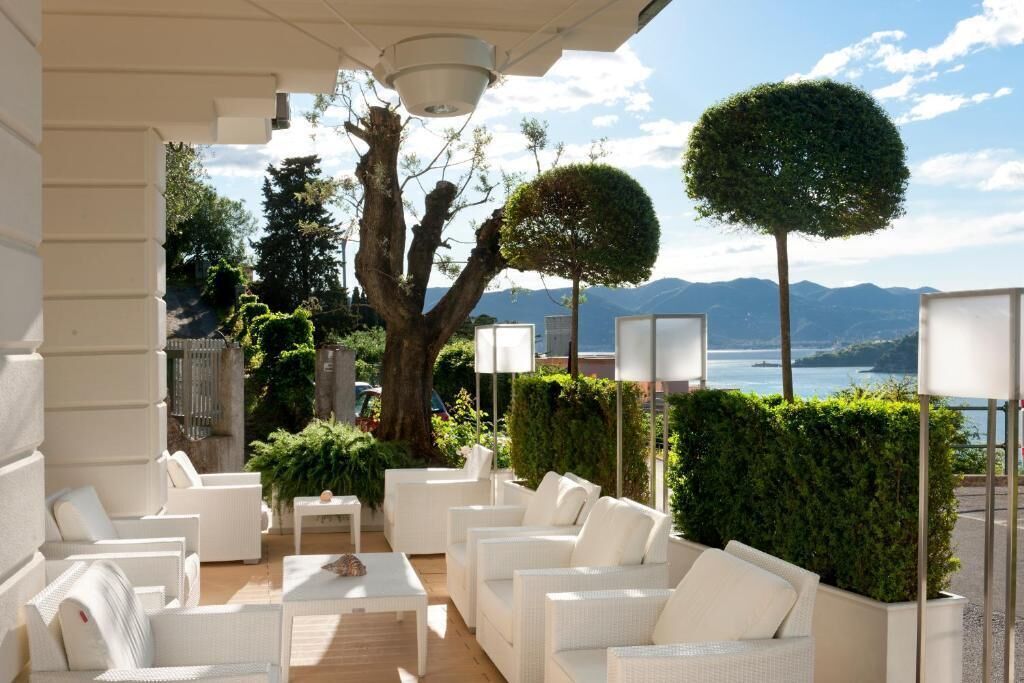 A terrace with white wicker furniture, a tree, and a view of a lake and mountains.