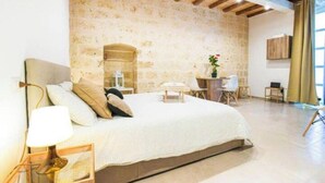 Een slaapkamer met een stenen muur, een bed met een witte spreidek, een nachtkastje met een lamp en een klein tafeltje met een bloevas.