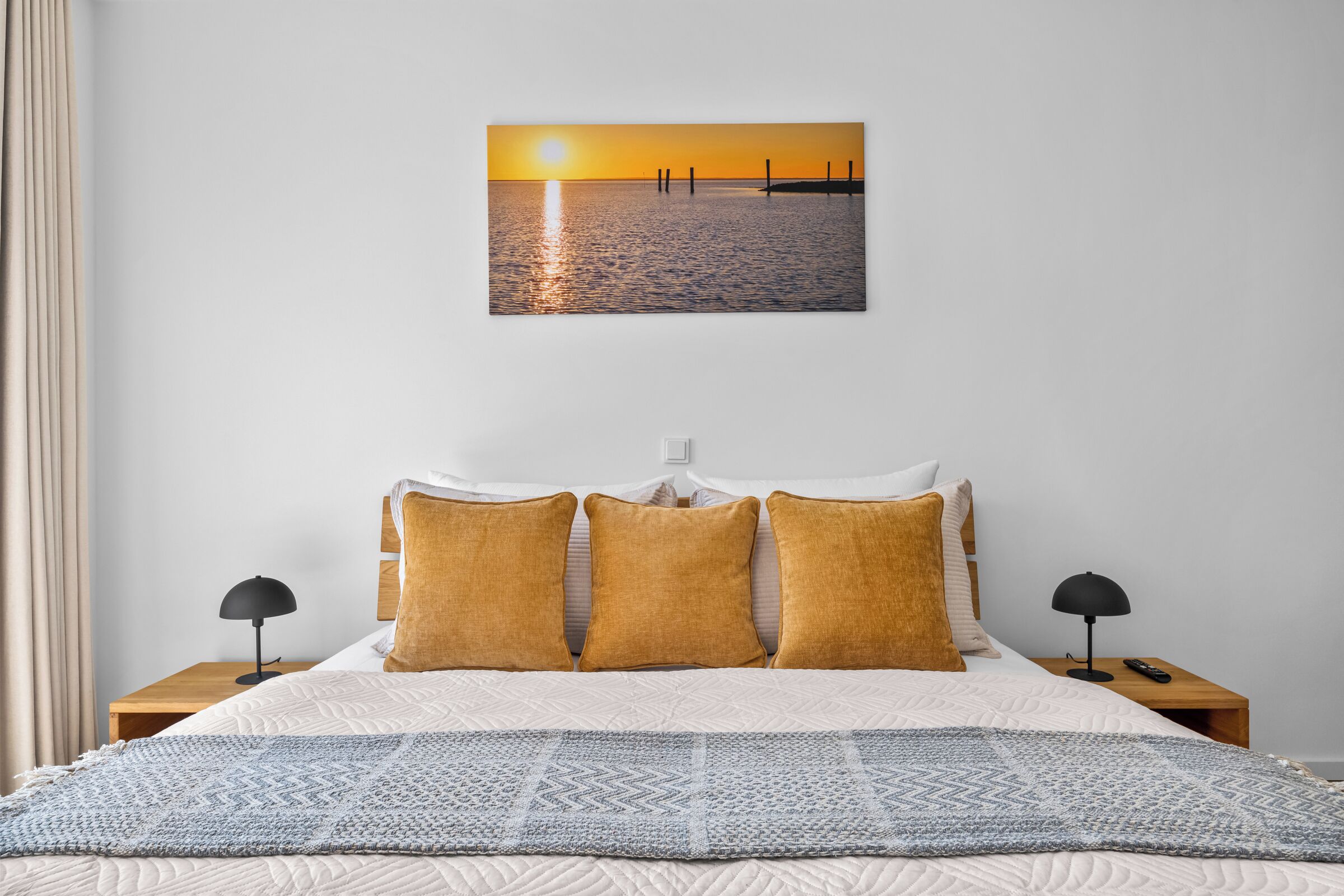 Une chambre d’hôtel avec un lit, deux tables de chevet, une petite table, une lampe et un tableau accroché au mur représentant un coucher de soleil sur l’eau.