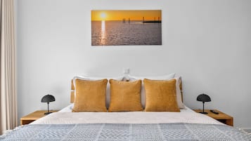 Une chambre d’hôtel avec un lit, deux tables de chevet, une petite table, une lampe et un tableau accroché au mur représentant un coucher de soleil sur l’eau.