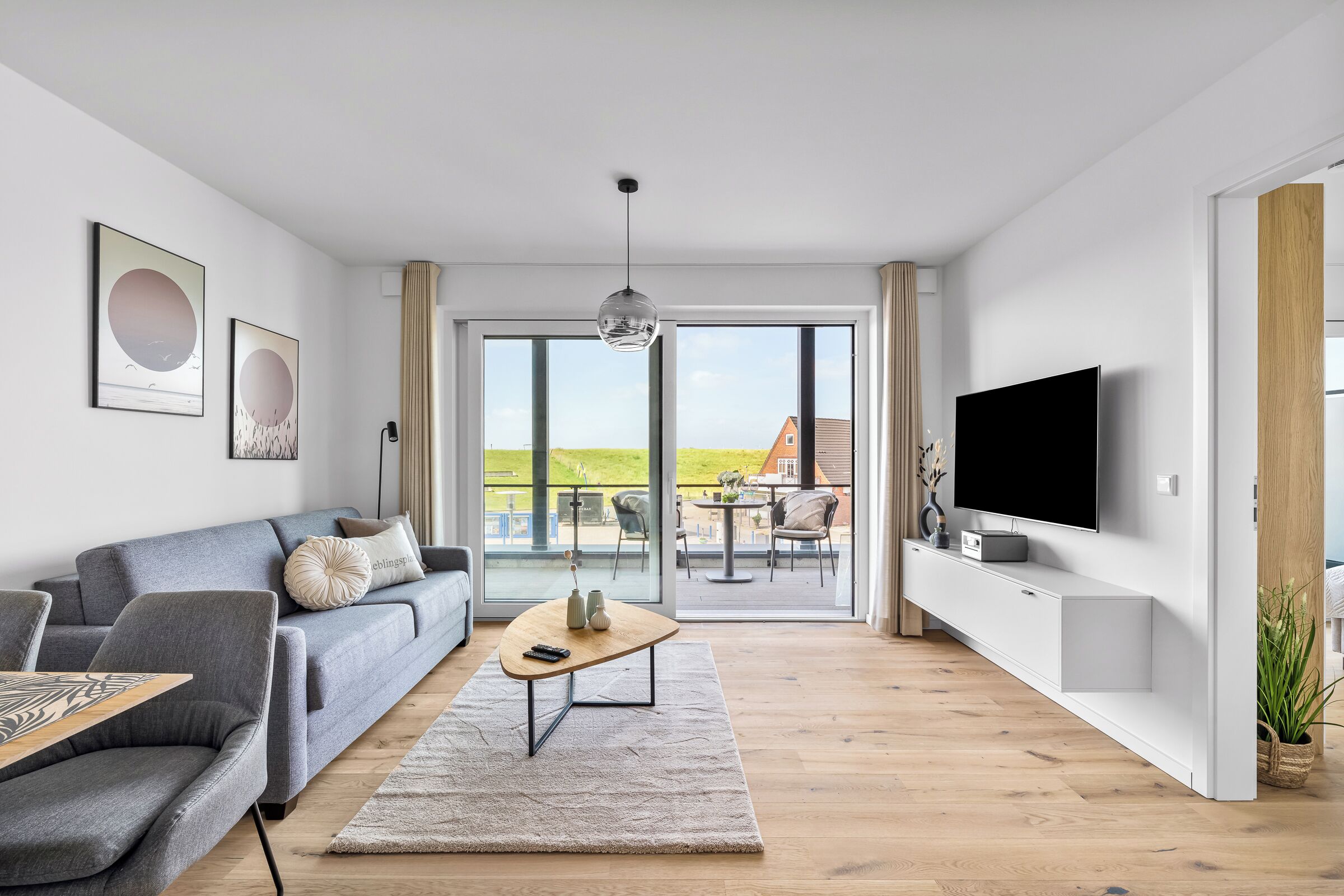 Ett modernt vardagsrum med en grå soffa, ett soffbord, en tv och ett stort fönster med utsikt över omgivningen.