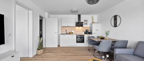 Un salon moderne avec un canapé gris, une table basse en bois et un téléviseur sur le côté gauche. On aperçoit à l’arrière-plan une cuisine avec des meubles blancs et un coin repas avec des chaises grises.