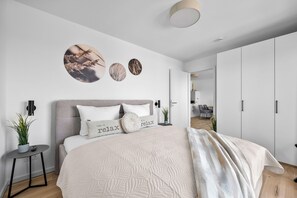 Ein modernes Schlafzimmer mit einem großen Bett, einem Nachttisch, einer Wandlampe und Blick in den angrenzenden Raum.