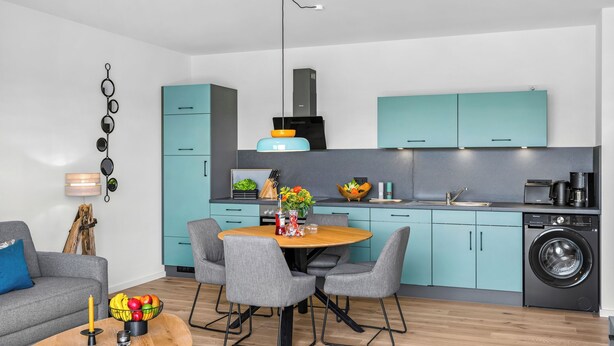 Una cocina moderna con gabinetes verde mar, lavadora integrada y un comedor con una mesa redonda y sillas grises.