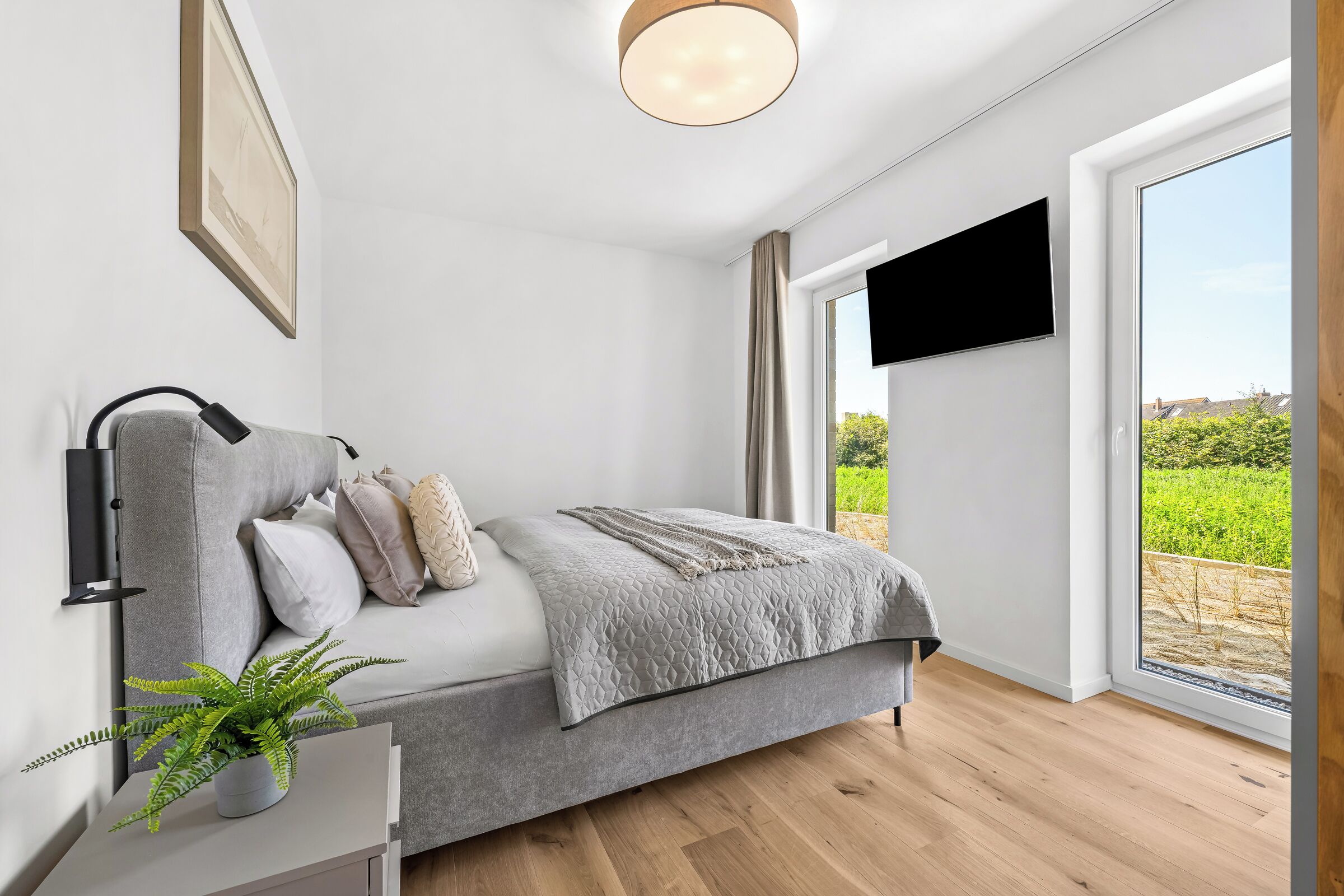 Un dormitorio moderno con cama, televisor montado, planta en maceta y un ventanal con vistas a zonas verdes.