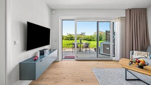 Una sala moderna con un televisor de pantalla plana, un mueble gris para equipo de entretenimiento, piso de madera, una puerta corrediza de vidrio que da a un patio exterior, y una zona de comedor con una mesa y sillas.