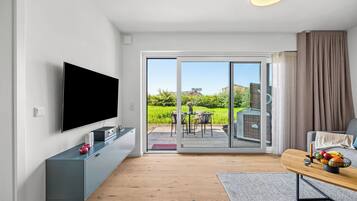 Ett modernt vardagsrum med en platt-TV, en grå möbel för underhållningssystemet, ett trägolv, en skjutdörr i glas som leder ut till en uteplats och en matplats med ett bord och stolar.